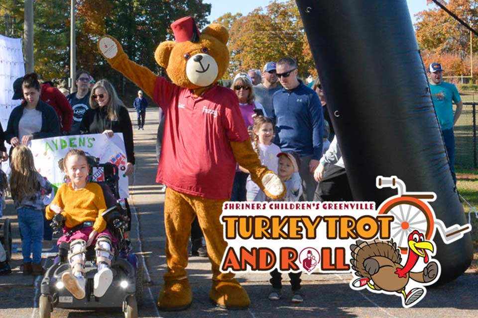 Patients, familles et autres participants, dont Fezzy, une grande mascotte d’ours, lors d’un événement précédent, et le logo de la Turkey Trot and Roll