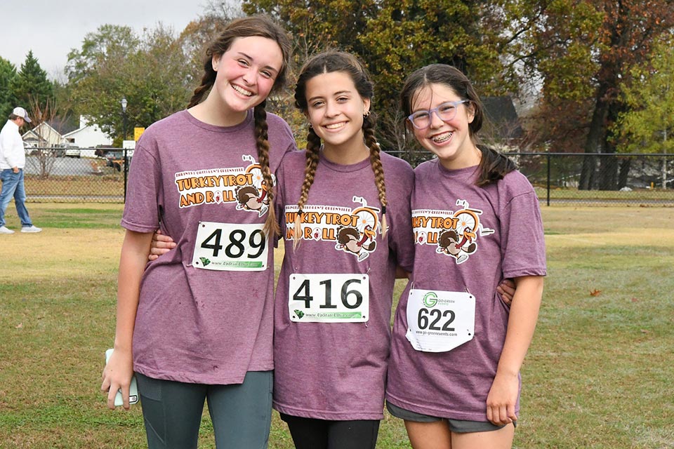Tres chicas sonrientes, con camisetas moradas de “Turkey Trot and Roll” haciendo juego y dorsales de carrera, tomadas del brazo en un campo con pasto.