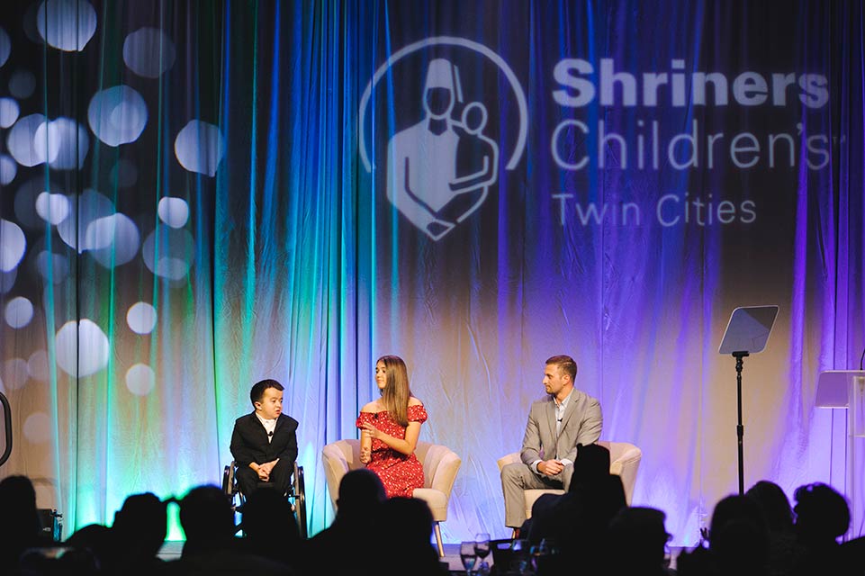 Alec, Gabbi y Parker en el escenario, el logotipo de Shriners Children's detrás de ellos
