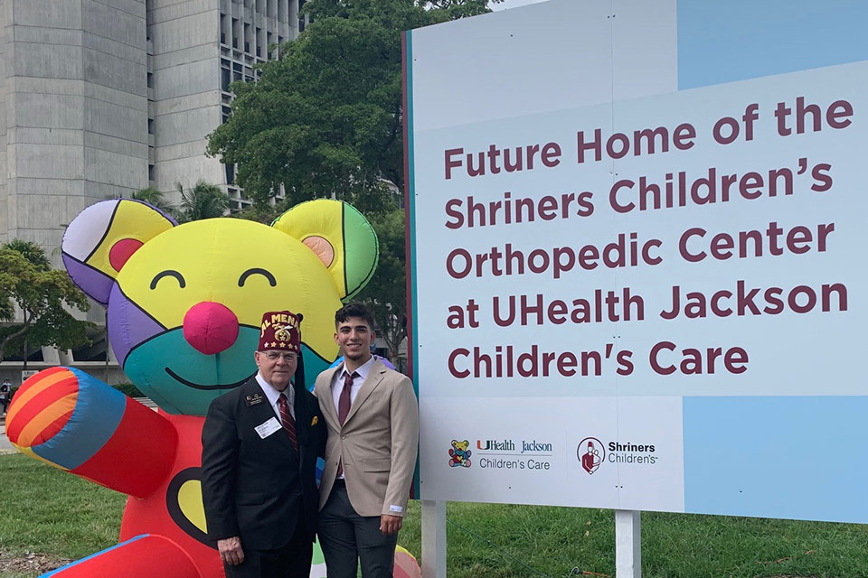 Patient à côté d’un Shriner et panneau indiquant « Future Home of the Shriners Children's Orthopaedic Center at UHealth Jackson Children's Care »