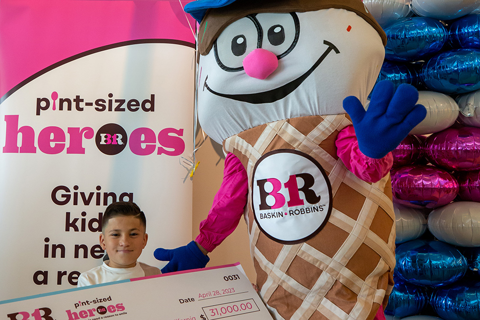 Sebastian avec la mascotte de Baskin-Robbins et un chèque géant de 31 000 $. L’arrière-plan montre des héros de la taille d’une pinte, donnant un aux enfants dans le besoin, logo Baskin-Robins.