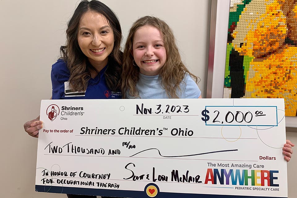 Courtney and therapist holding fake check, language on check: Nov. 2, 2023, Shriners Children's Ohio logo, $2,000, In honor of Courtney for occuplational therapy, signatures, the most amazing care anywhere, pediatric specialty care