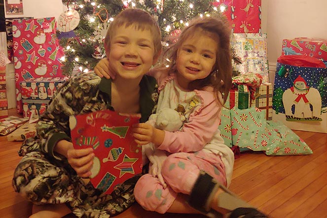 Scarlett con su hermano abriendo regalos con un árbol de Navidad en segundo plano
