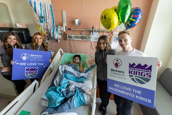 Emmy visits patients at the hospital, signs with text reading "We love the Sacramento Kings," Shriners Children's logo, Sacramento Kings logo