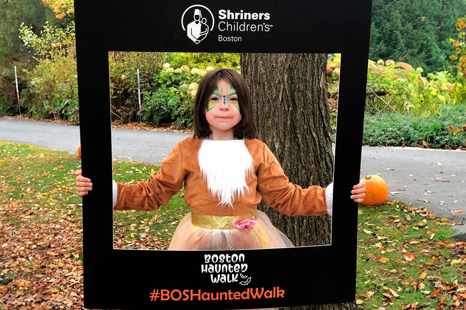 Emily sosteniendo un marco, logotipo del Hospital Shriners para Niños Boston, logotipo del paseo embrujado de Boston, #BOSHauntedWalk