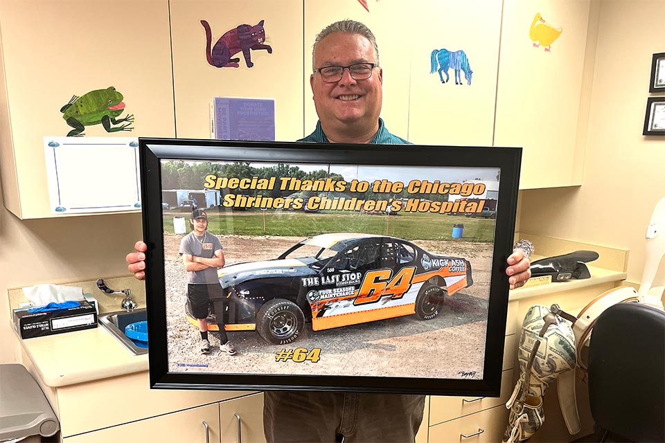 provider holding framed photo; framed photo: race car, image headline: Special Thanks to the Chicago ShrinersChildren's Hospital; car wrap language: Kick Ash Coffee, Four Seasons Maintenance, The Last Stop. Brady in photo.