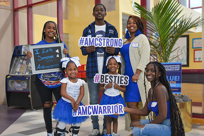 MiKenli con cinco miembros de su familia sosteniendo carteles que decían: Amo a alguien con artrogriposis, AMC Strong, Besties, AMC Warrior