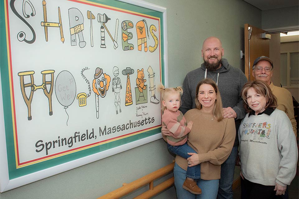 Expaciente con su familia frente al cartel del Hospital Shriners para Niños de New England