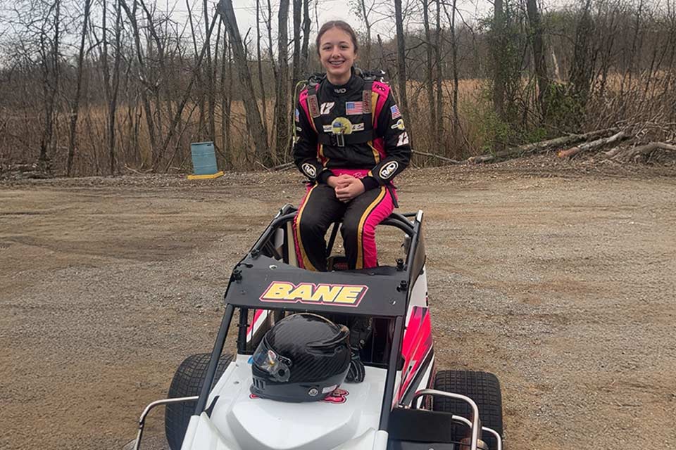 a young racecar driver sitting on top of her car, which features the word "BANE" written across the top