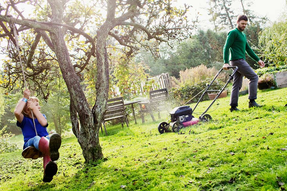 Père tondant la pelouse pendant que sa fille, sur une balançoire, le regarde