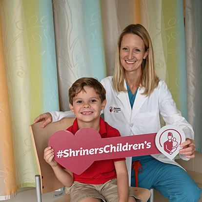 Female doctor sitting next to young male holding a Shriners Children's sign
