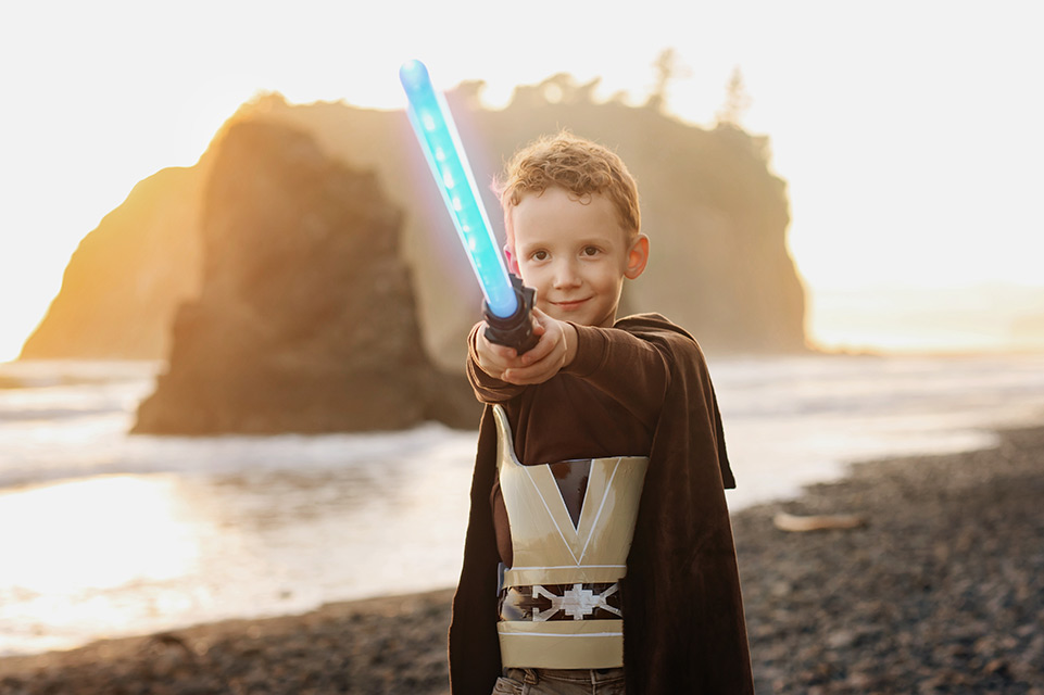 Petit garçon debout sur une plage, costumé, tenant une épée lumineuse en plastique